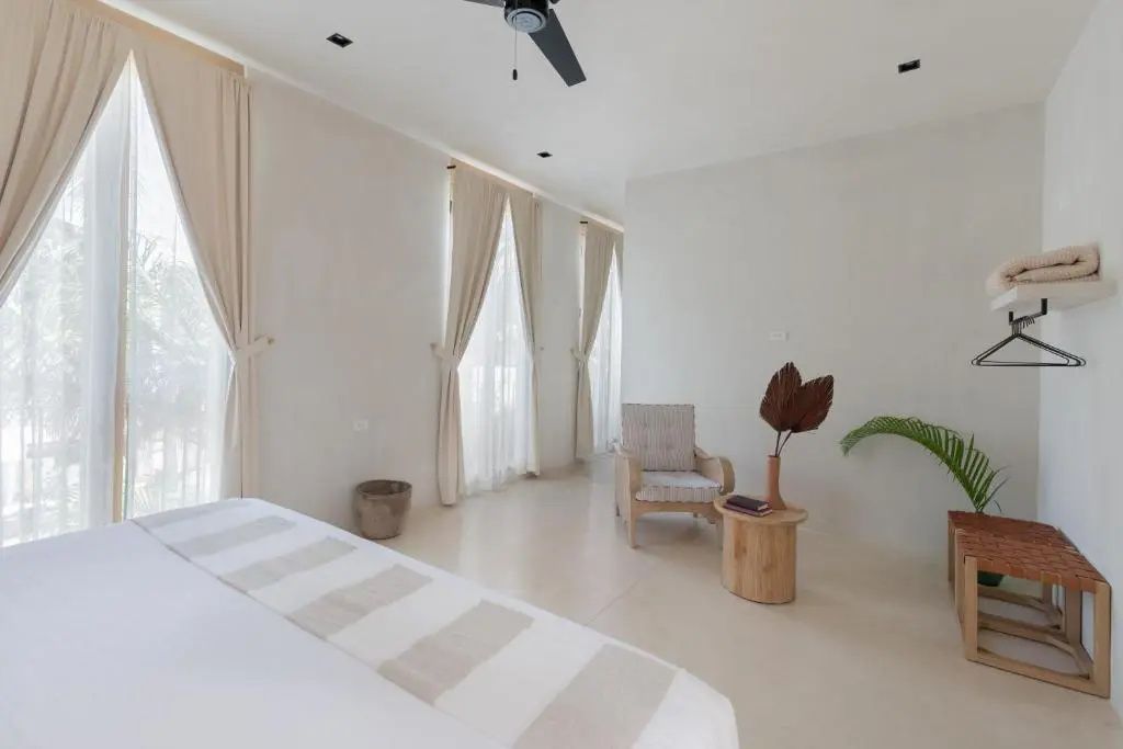 Helles Schlafzimmer mit beigen Vorhängen, Holzsessel und kleiner Sitzecke.