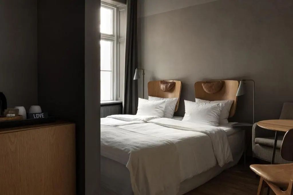 Hotelzimmer mit einem weißen Doppelbett, Wandleuchten und einem Fenster mit dunklem Vorhang.