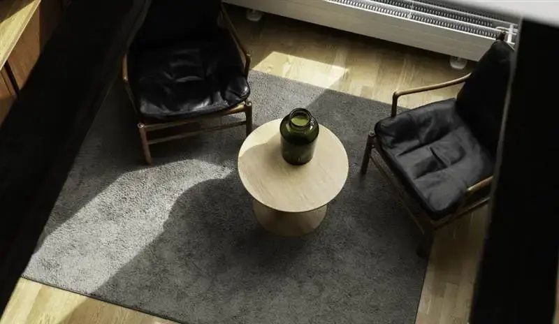 Runder Holztisch mit grüner Glasvase zwischen zwei schwarzen Ledersesseln auf einem grauen Teppich.