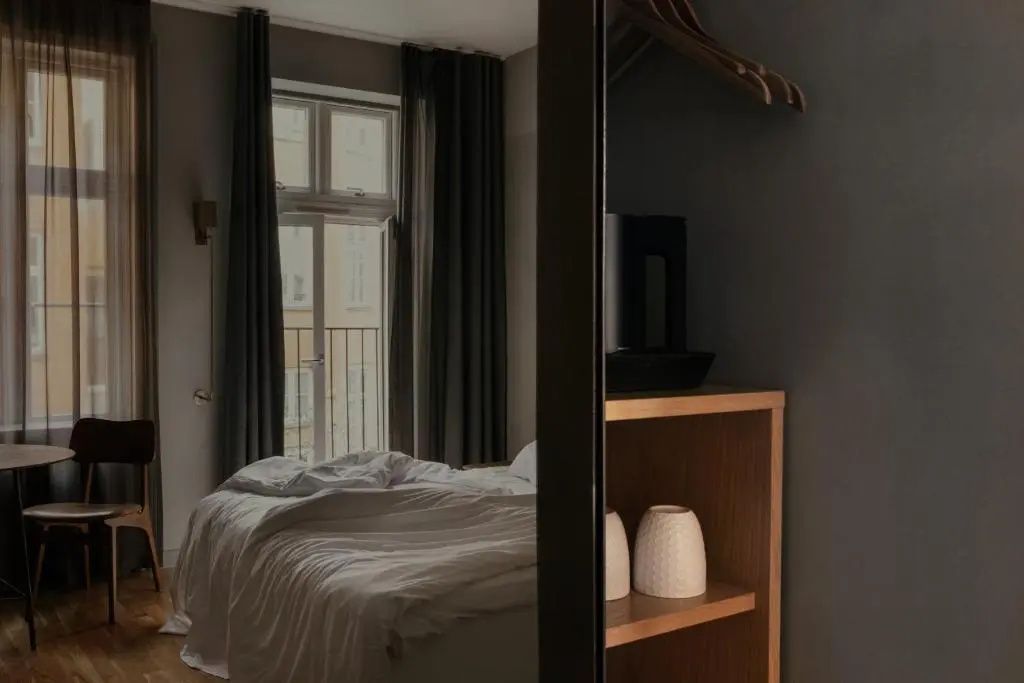 Blick an einem Holzregal vorbei auf ein Bett vor dem Fenster mit langen Vorhängen.