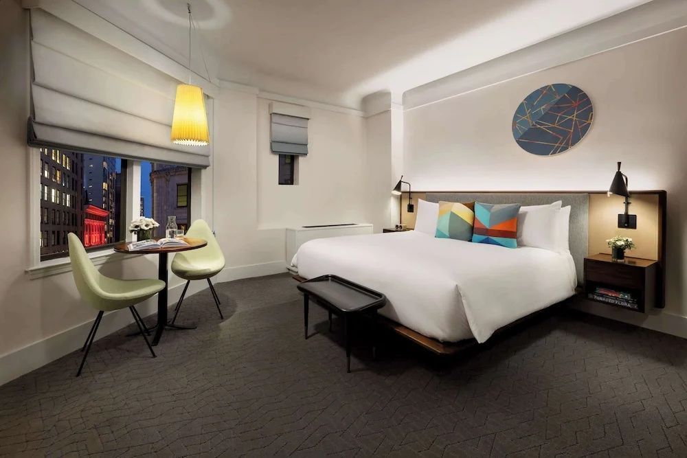 Modernes Hotelzimmer mit großem Doppelbett, bunten Kissen und einem kleinen Tisch mit zwei Stühlen.