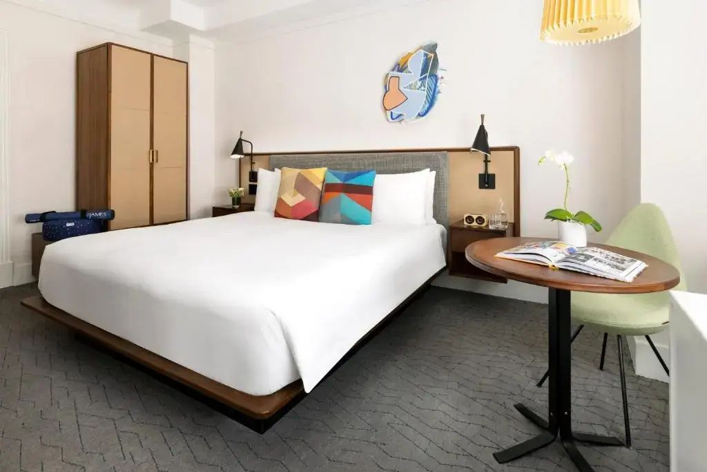 Doppelbett mit bunten Kissen, Wandkunst und einem kleinen Tisch im hellen Hotelzimmer.