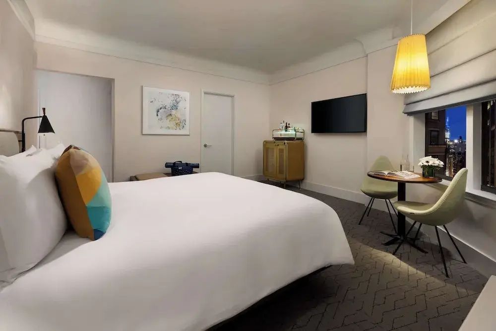Blick auf ein Hotelzimmer mit Flachbildfernseher an der Wand, Kommode und gelber Hängelampe.