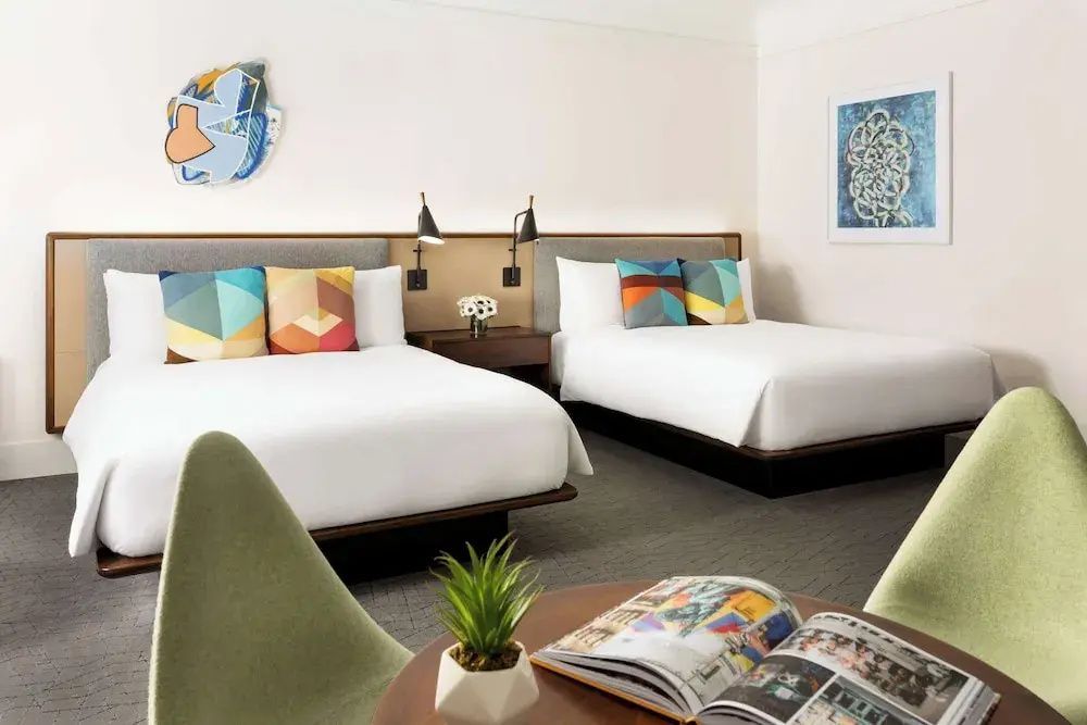 Geräumiges Hotelzimmer mit zwei Doppelbetten, bunten Kissen und Kunst an den Wänden.