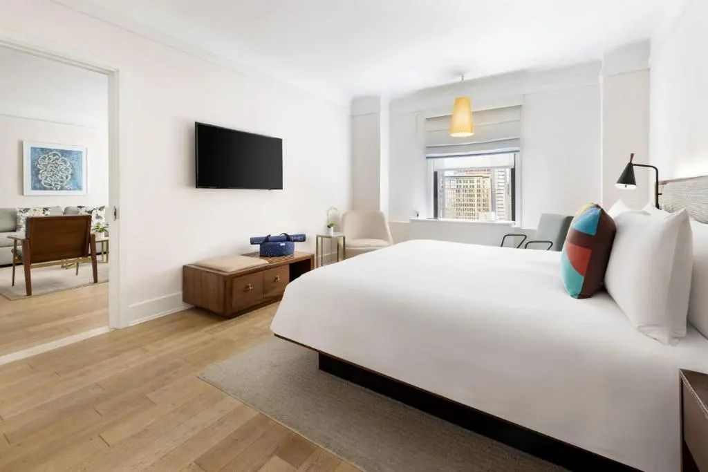 Modernes Hotelzimmer mit einem Doppelbett, Holzboden, Fernseher und Sessel am hellen Fenster.