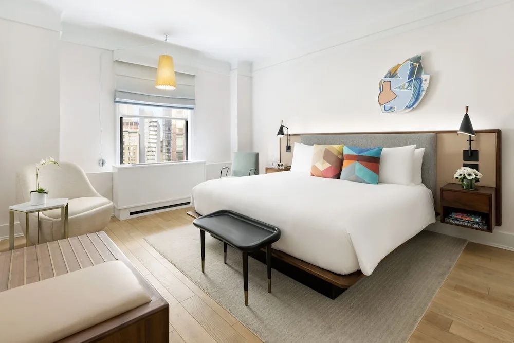 Helles Hotelzimmer mit großem Bett, bunten Kissen, Holzfußboden und Fenster zur Stadt.