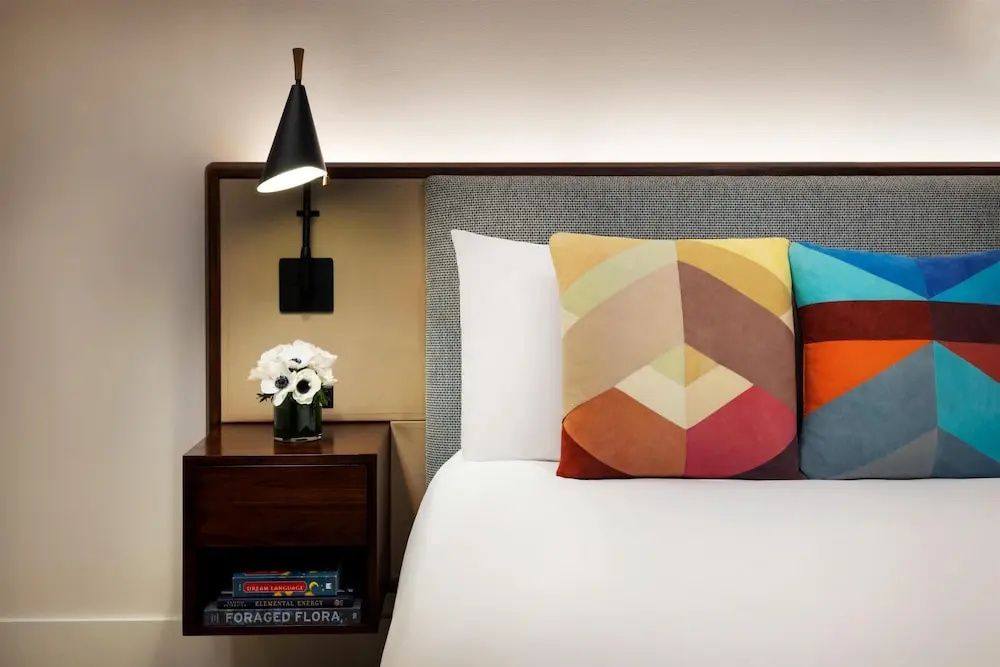 Nachttisch aus Holz mit kleiner Lampe, Blumenstrauß, Büchern und buntem Kissen auf dem Bett.