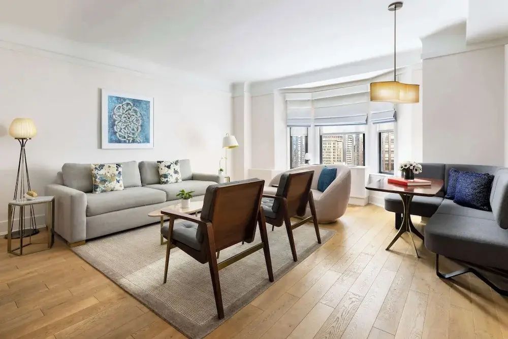 Wohnzimmer einer Suite mit grauem Sofa, Sesseln aus Holz und großen Fenstern mit Stadtblick.