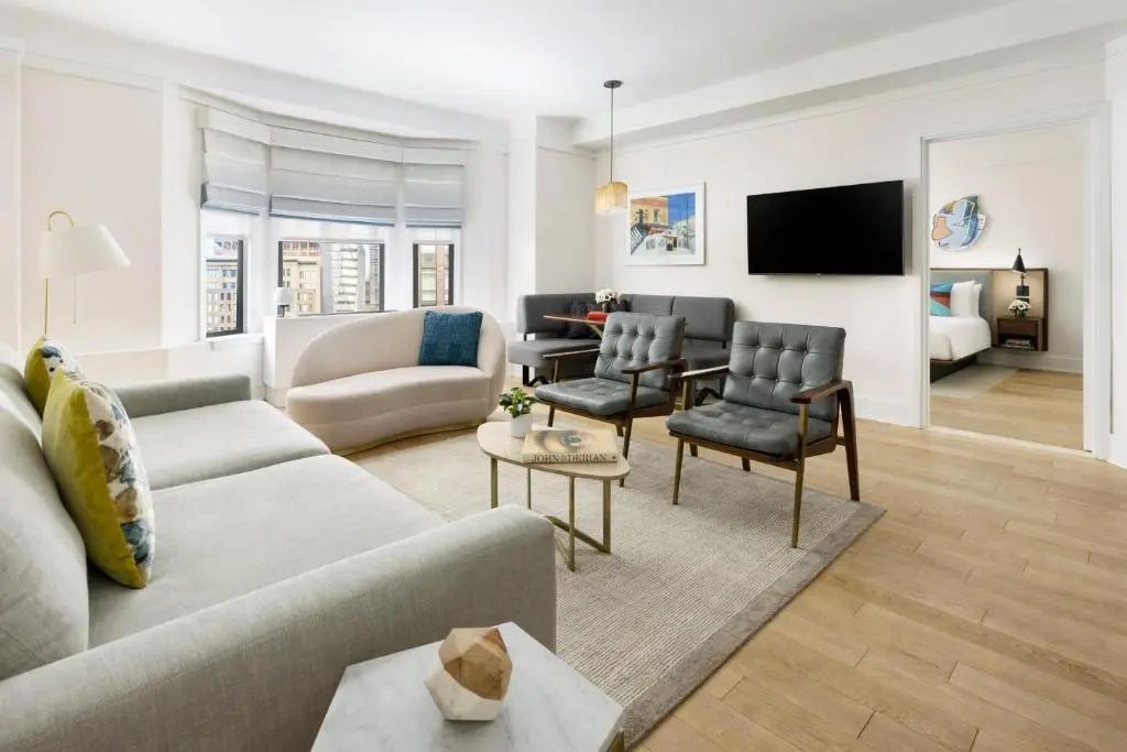 Wohnbereich mit hellem Sofa, zwei grau gepolsterten Sesseln, Fernseher und Erkerfenster.