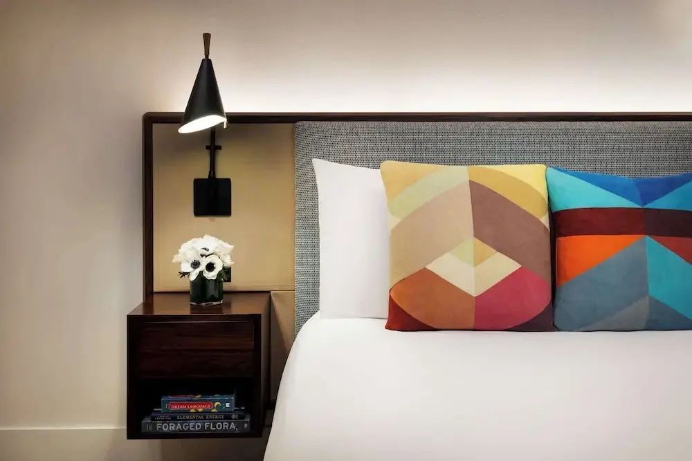Nachttisch mit einer schwarzen Lampe, einem kleinen Blumenstrauß und geometrisch gemusterten Kissen.