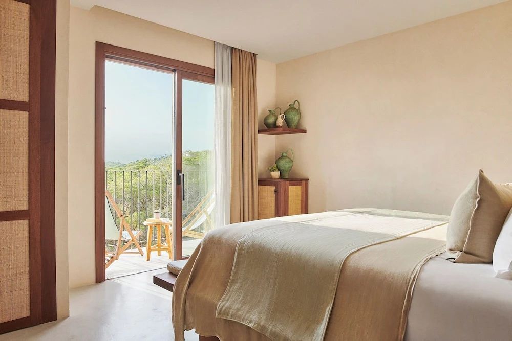 Helles Schlafzimmer mit Bett und geöffneter Balkontür mit Blick in die Natur.