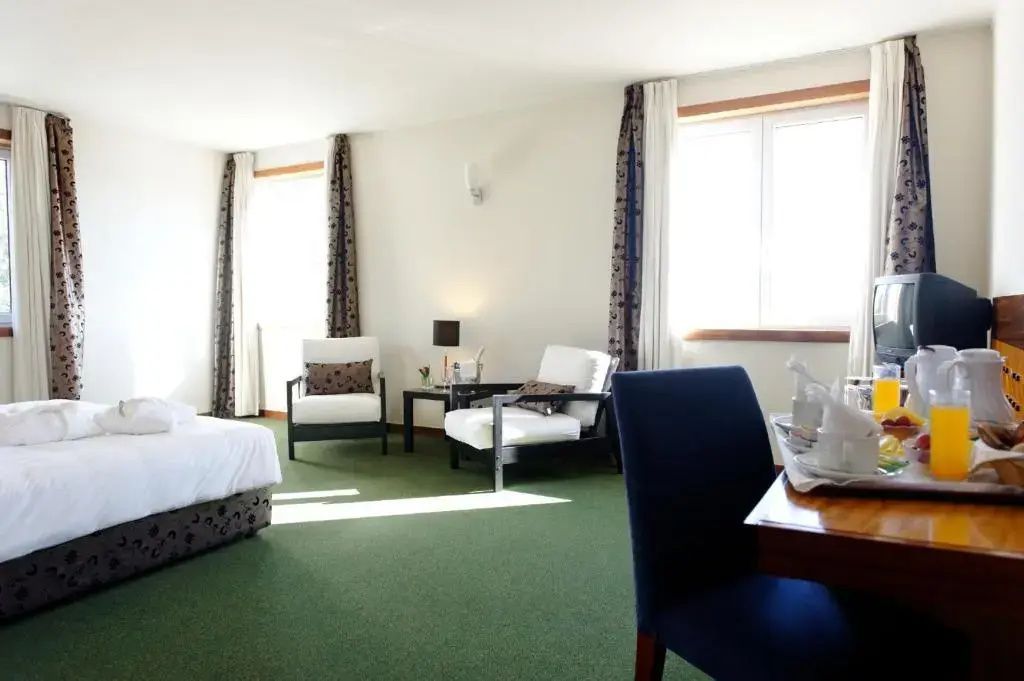 Helles Hotelzimmer mit grünem Teppichboden, zwei weißen Sesseln und großen Fenstern.