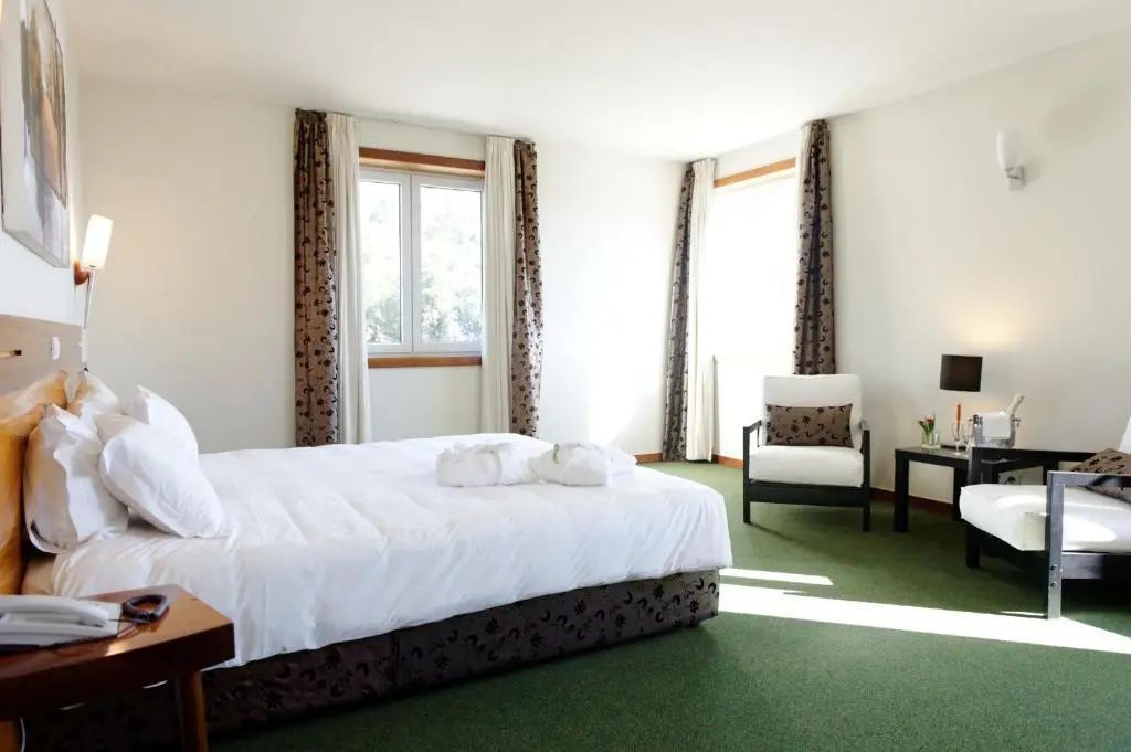 Helles Hotelzimmer mit Doppelbett, grünem Teppichboden und einem Sessel am Fenster.