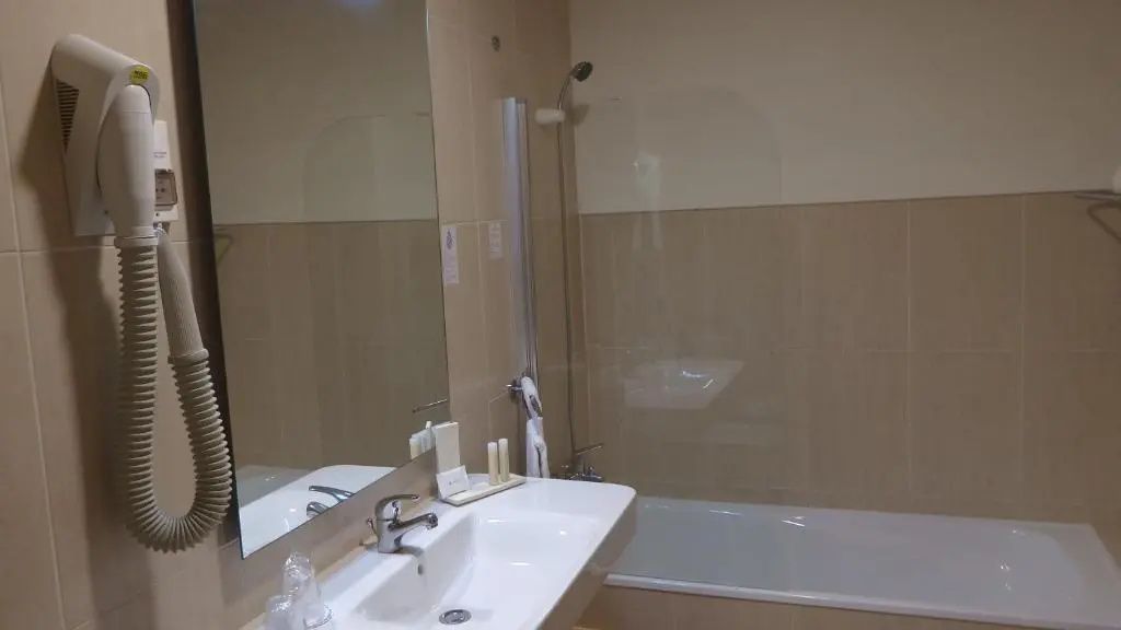 Badezimmer im Hotel mit weißem Waschbecken, großem Spiegel und einer Badewanne mit Glasabtrennung.