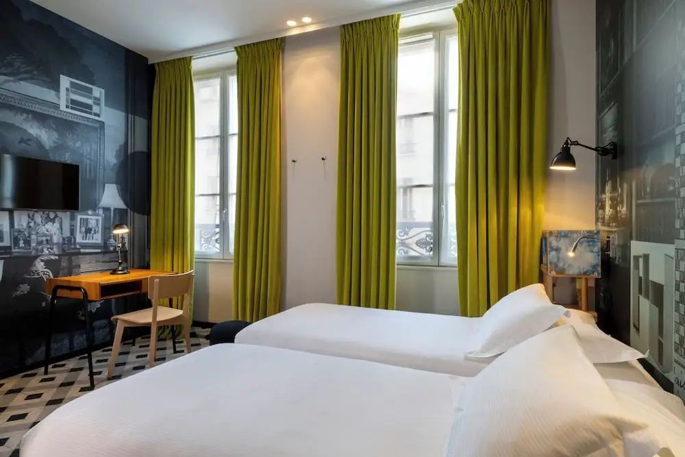 Zweibettzimmer mit zwei weiß bezogenen Betten, grünen Vorhängen und kleinem Schreibtisch.