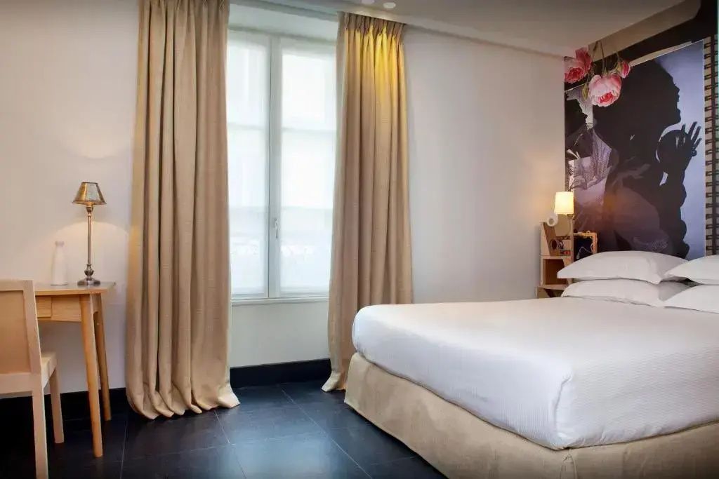 Doppelbett mit weißer Bettwäsche neben hohen Vorhängen und einem hellen Fenster.