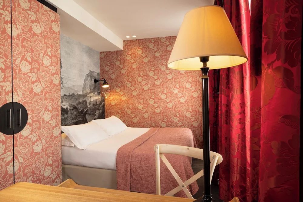 Hotelzimmer mit roter gemusterter Tapete, einem Doppelbett und einer großen Stehleuchte.