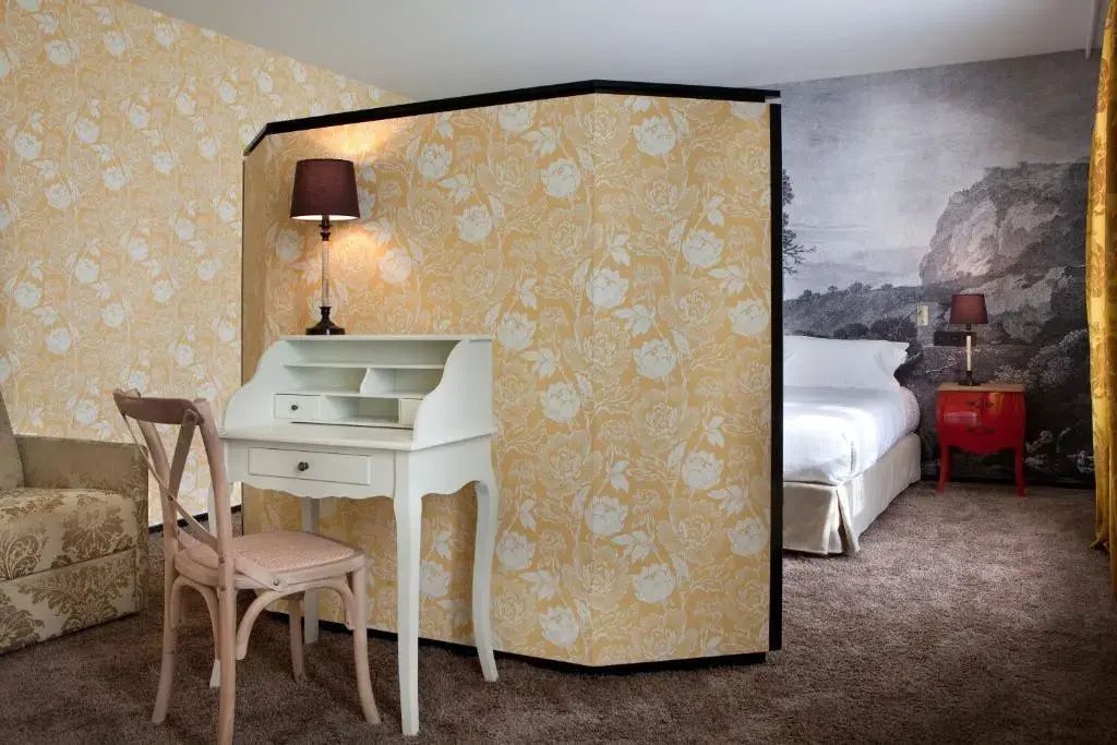 Hotelzimmer mit gelb gemusterter Trennwand, kleinem Schreibtisch und Blick zum Bett.