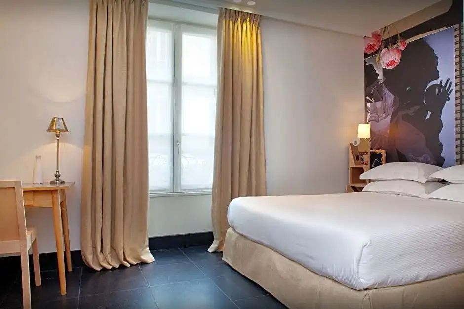 Helles Hotelzimmer mit großem Doppelbett, beigen Vorhängen und dunklem Fliesenboden.