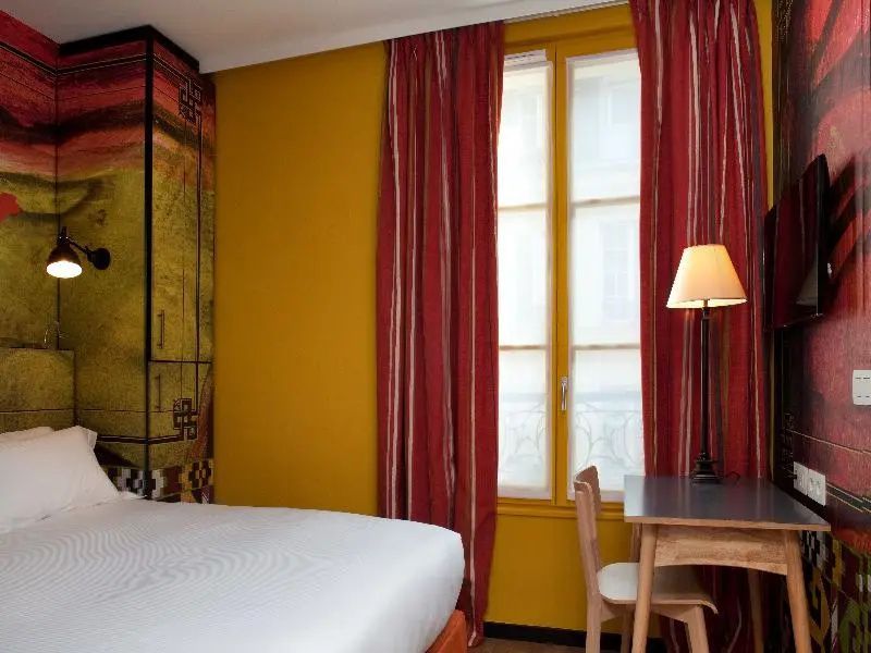 Hotelzimmer mit gelben Wänden, roten Vorhängen am Fenster und einer kleinen Arbeitsecke.