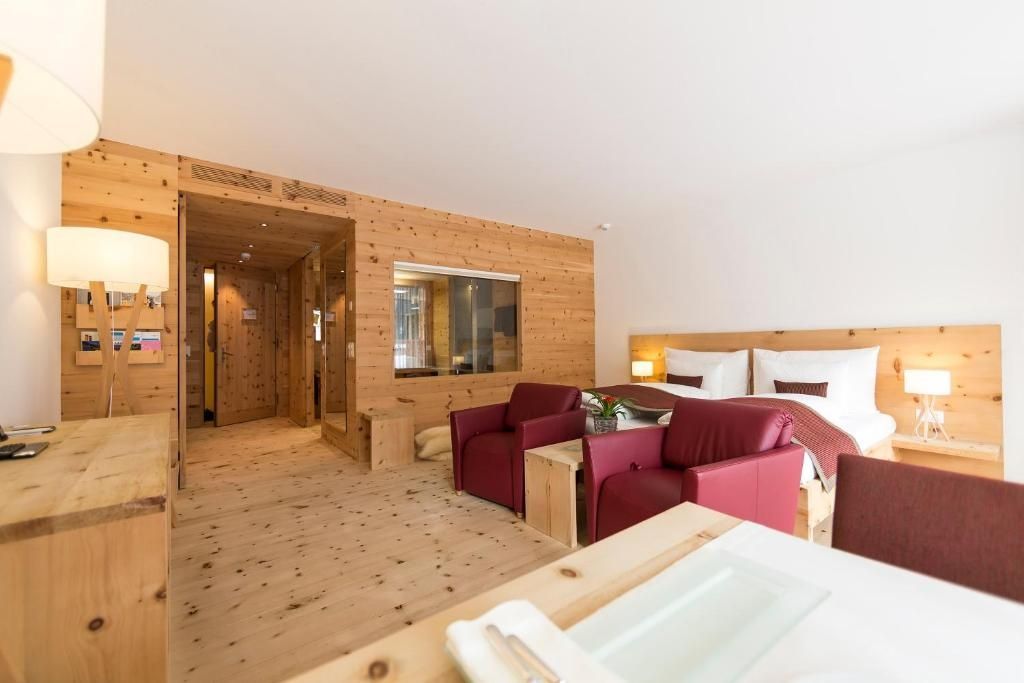 Hotelzimmer aus Arvenholz mit zwei roten Sesseln, Tisch und Blick zum Schlafbereich.
