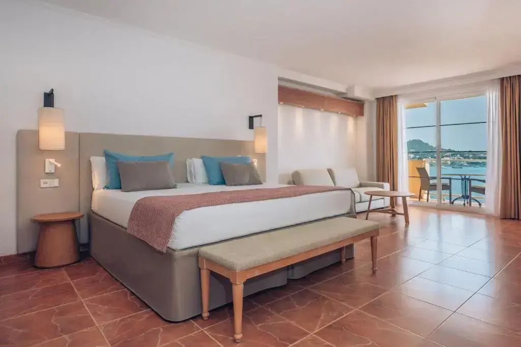 Helles Hotelzimmer mit großem Doppelbett, Sitzbank, Sofa und Balkonzugang mit Meerblick.