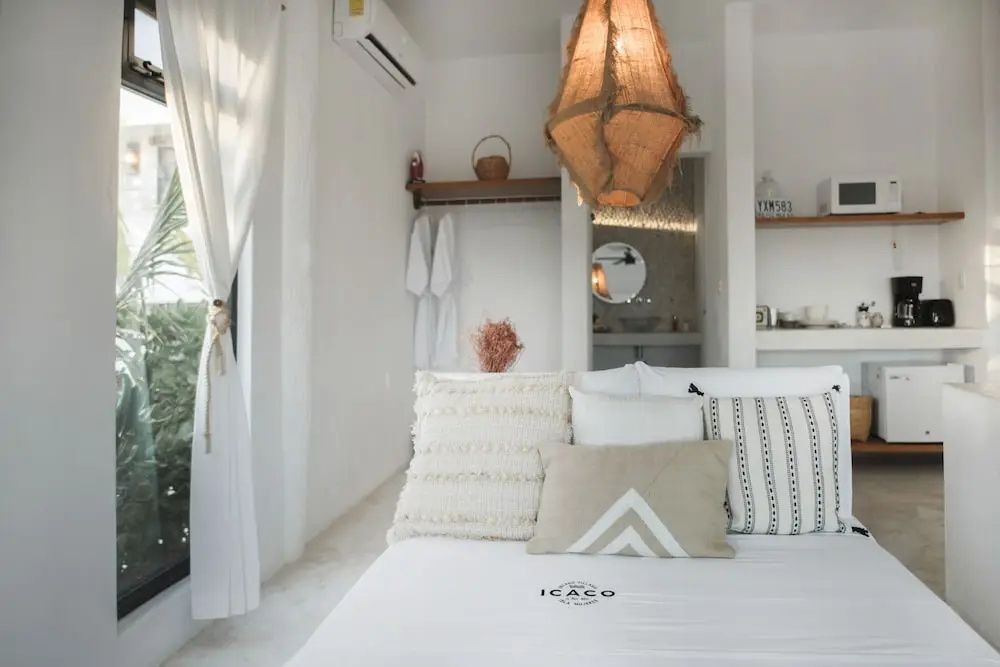 Bett mit weißer Bettwäsche und Kissen in einem weiß gestalteten Zimmer mit Hängelampe.