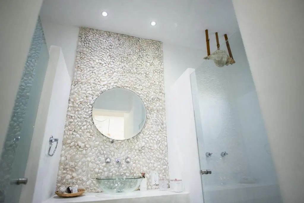 Badezimmer mit steinbesetzter Wand hinter dem rundem Spiegel und einer Dusche aus Glas.