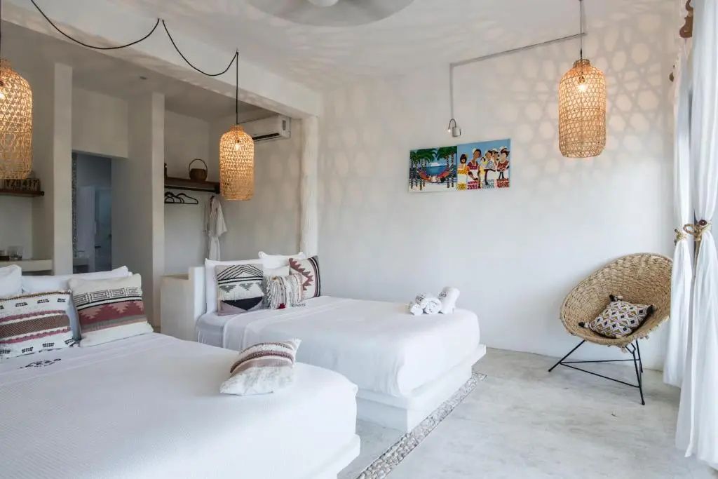 Geräumiges Hotelzimmer mit zwei weißen Betten, Flechtlampen und bunten Wandbildern auf weißer Wand.
