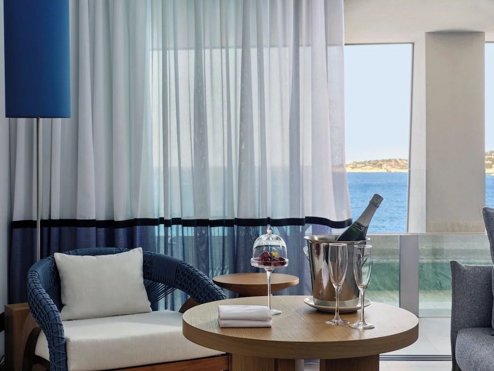 Sitzbereich im Hotelzimmer mit Holztisch, Sektkühler und Blick auf das blaue Meer.