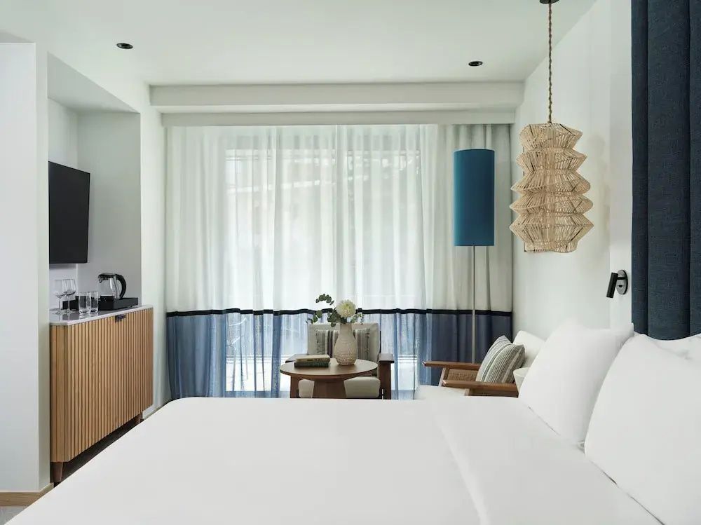 Hell eingerichtetes Hotelzimmer mit großem Doppelbett, Sitzecke und Vorhängen.