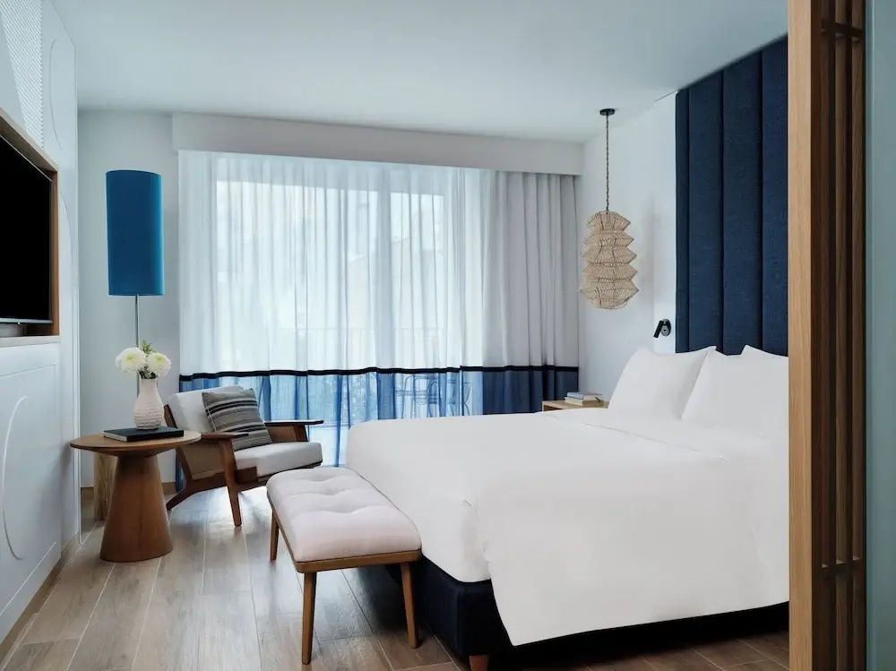 Doppelbett mit weißer Bettwäsche, Sitzbank und Vorhängen vor der Fensterfront.