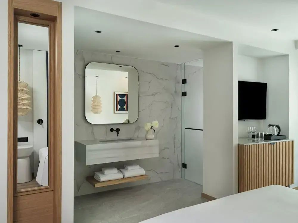 Offen gestaltetes Hotelzimmer mit modernem Waschtisch, Wandspiegel und Fernseher.