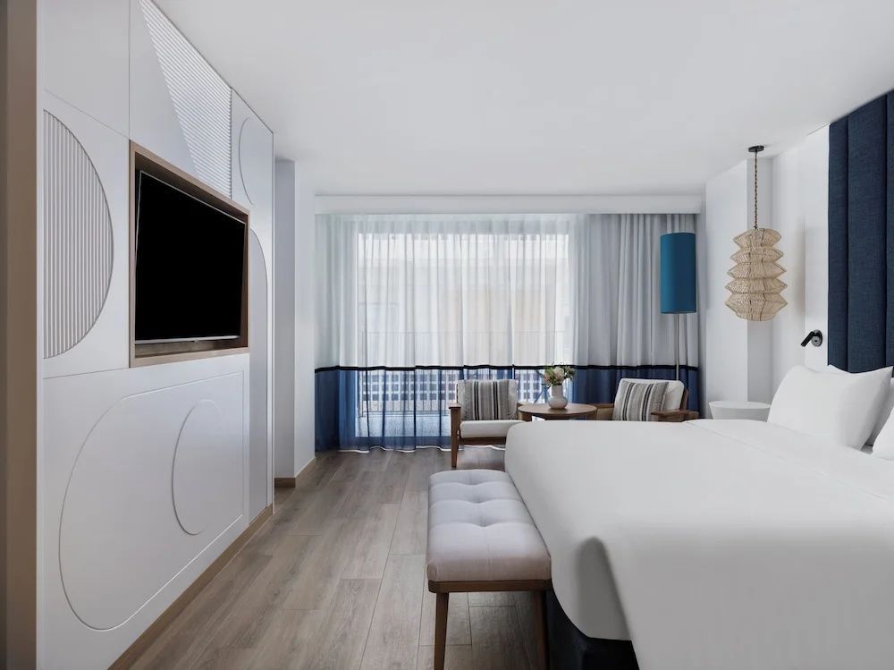 Helles Hotelzimmer mit großem Doppelbett, Sitzbereich vor dem Fenster und Wandfernseher.