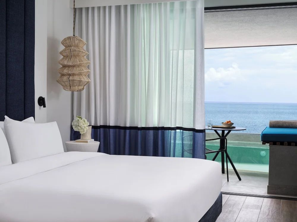 Blick vom Bett durch transparente Vorhänge auf einen Balkontisch und das blaue Meer.