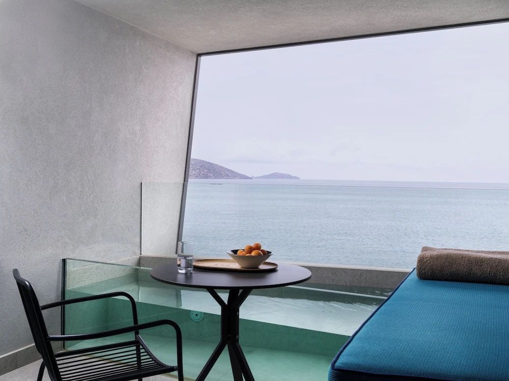 Balkon mit kleinem Tisch, Liegemöglichkeit und Glasgeländer vor weitläufiger Meereskulisse.