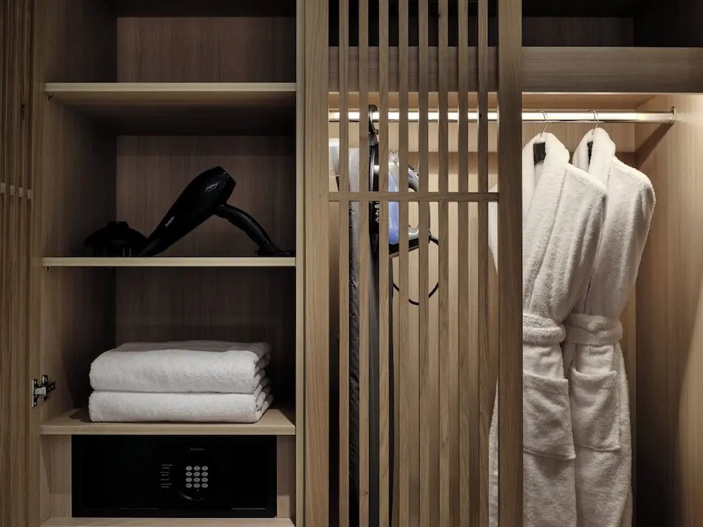 Offener Kleiderschrank aus Holz mit Safefach, Föhn und weißen Bademänteln.