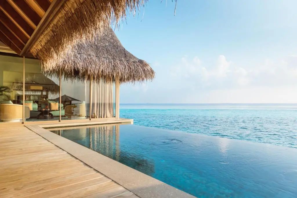 Infinity-Pool einer Wasser-Villa mit Reetdach direkt am blauen Ozean unter klarem Himmel.