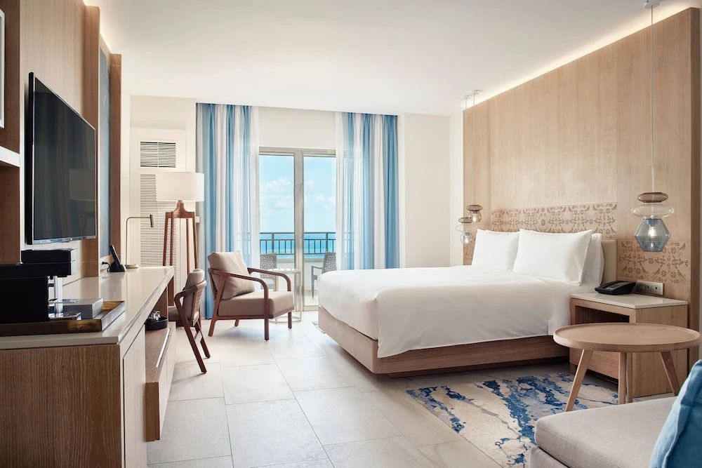 Helles Hotelzimmer mit großem Doppelbett, Sessel und Balkontür mit Blick auf das Meer.