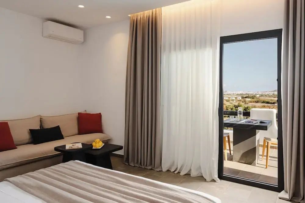Helles Hotelzimmer mit Bett, beigem Sofa mit rotem Kissen und großem Fenster zum Balkon.