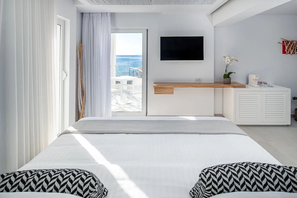 Modernes helles Hotelzimmer mit Doppelbett, Fernseher an der Wand und Meerblick durch die Balkontür.