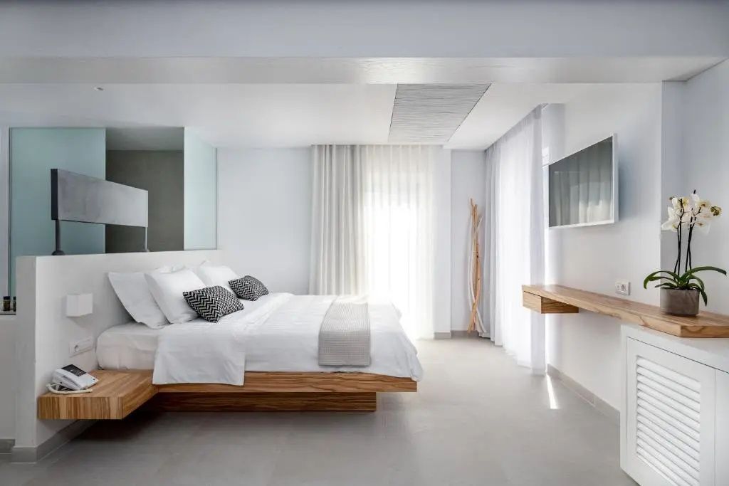 Modernes Hotelzimmer in Weißtönen mit großem Doppelbett und Holzgestell sowie Wandfernseher