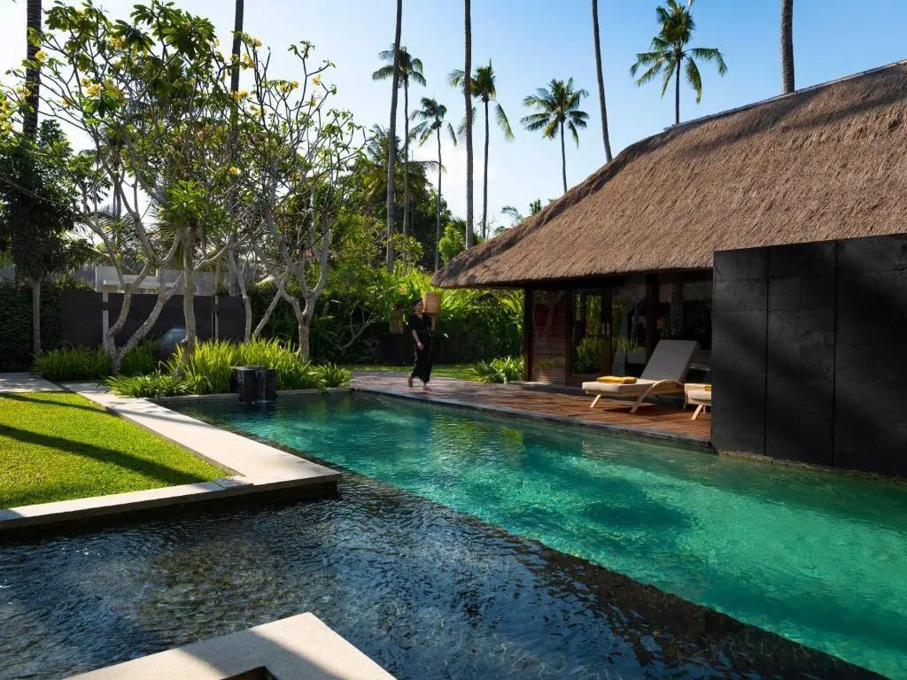 Türkisfarbener Pool im Garten neben einer Villa mit Reetdach und hohen Palmen.