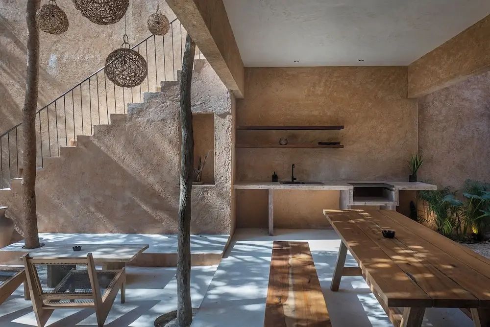 Offener Außenbereich mit Holztisch, Holzbank, Treppe und einer Wand in Naturtönen.