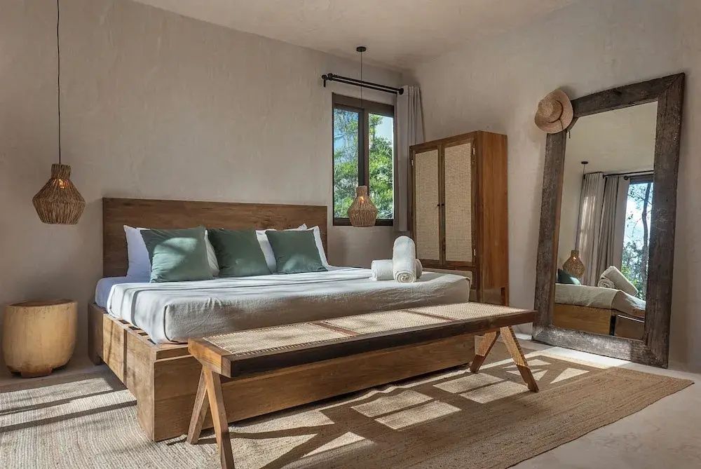Schlafzimmer mit großem Holzdoppelbett, grünen Kissen, Holzbank und einem hohen Spiegel.