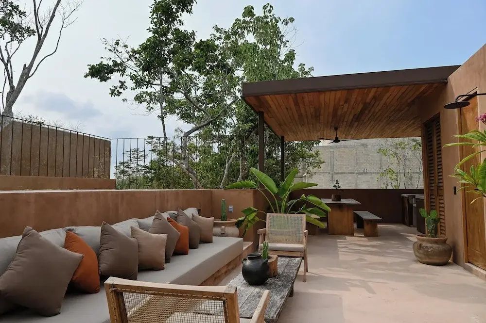 Dachterrasse mit langer Sitzbank, Kissen, Holztisch unter einer Überdachung und Blick auf Bäume.