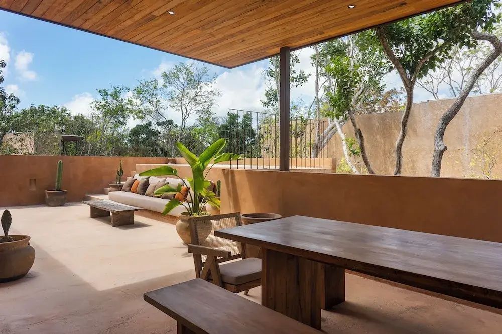 Überdachte Terrasse mit Holzmöbeln, Sitzbank und Blick auf umliegende Bäume.