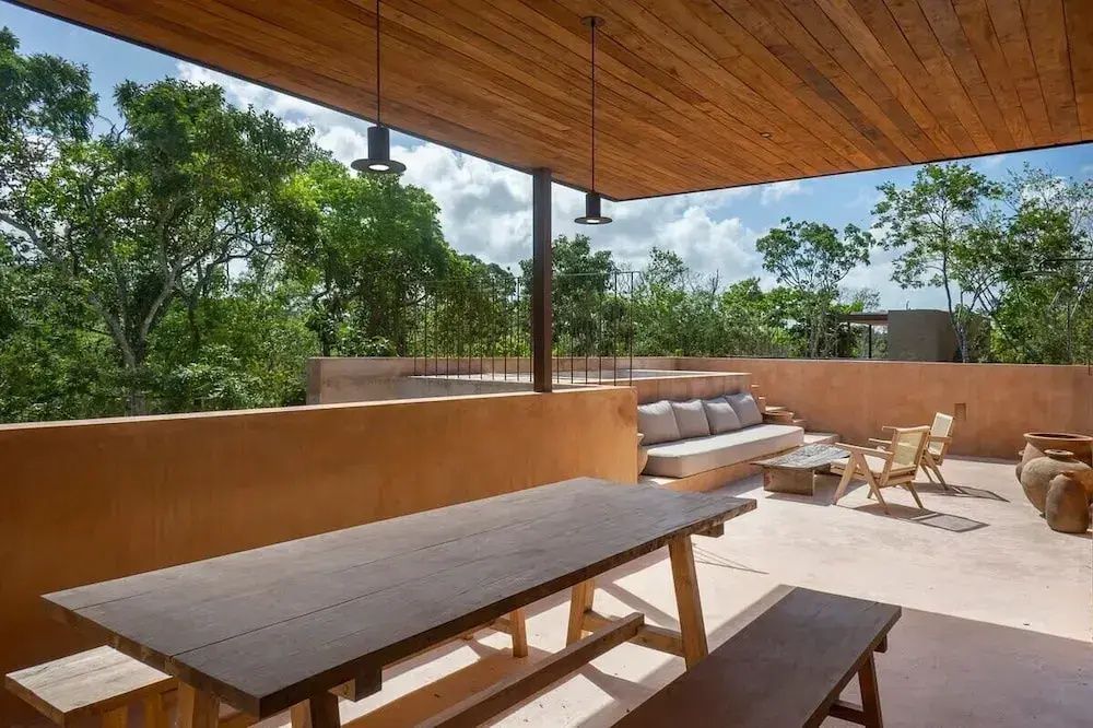 Holztisch und Sitzbank auf einer Dachterrasse mit Blick auf tropische Bäume.