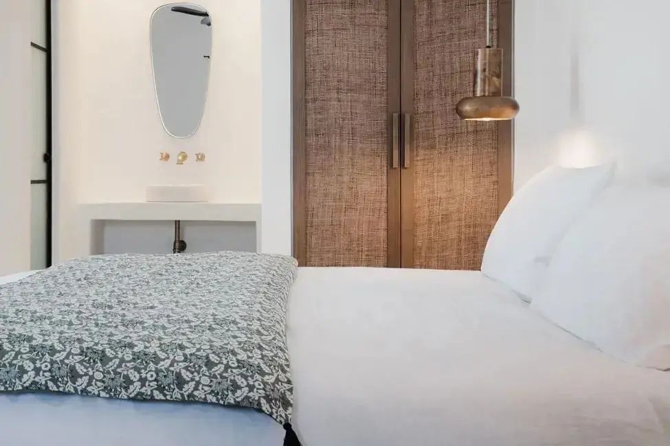 Helles Hotelzimmer mit weiß bezogenem Bett, gewebtem Schrank und Wandspiegel im Hintergrund.