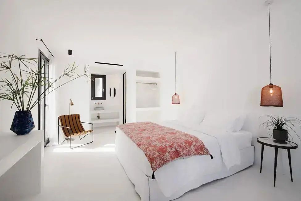Helles Hotelzimmer mit Doppelbett, rosa Tagesdecke, Sessel und Blick ins Badezimmer.